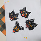 Four die-cut vinyl stickers featuring stylized black animal heads with orange and yellow eyes and red accents, arranged on a white sheet of paper.