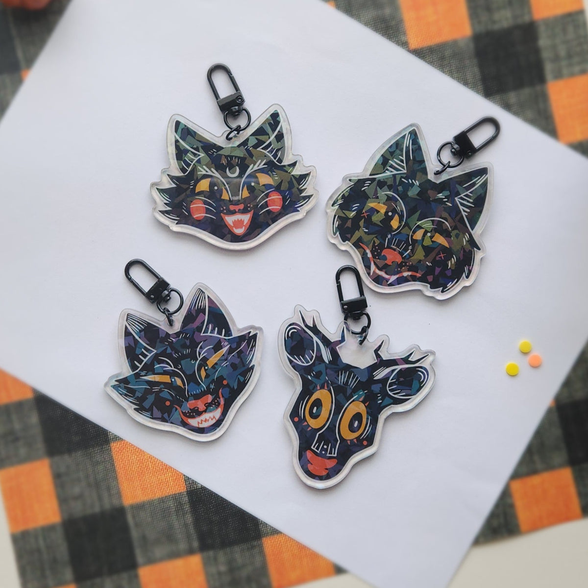 Four stylized animal head keychains with black carabiners, arranged on a white sheet of paper over an orange and black plaid fabric background.