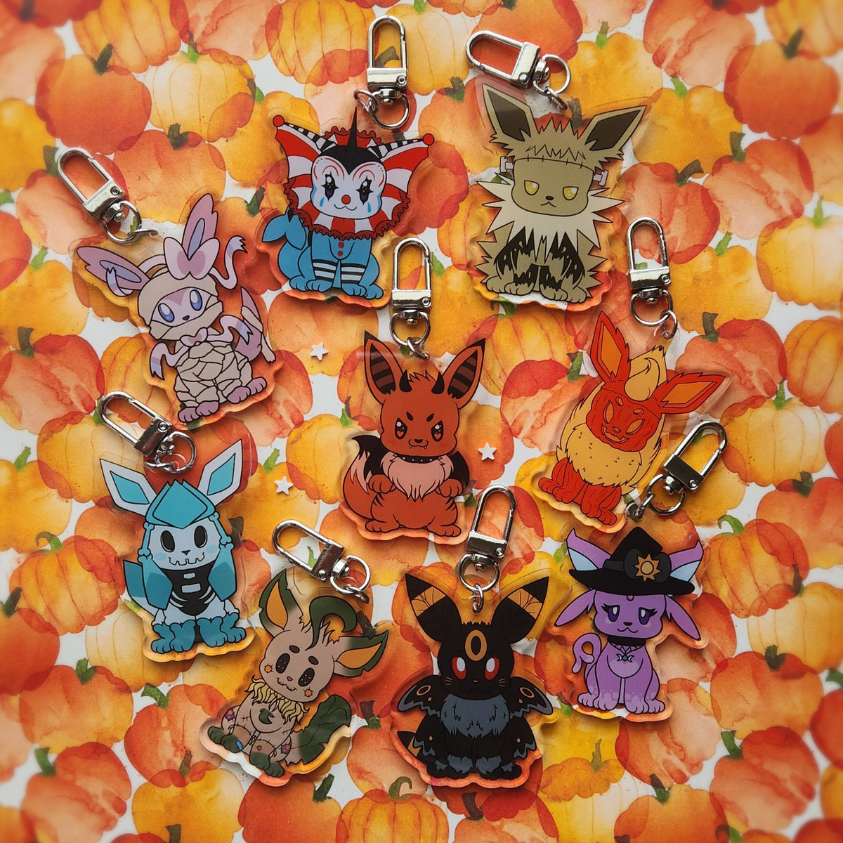 Nine colorful acrylic keychain charms shaped like different Halloween foxes, arranged on a pumpkin-patterned background. Each with silver metal keychain clasp attachments.