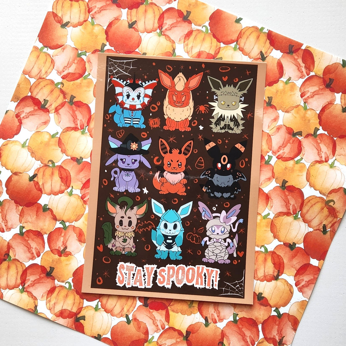 A rectangular Halloween greeting card featuring cute cartoon foxes on a dark brown background with the words 'STAY SPOOKY!' at the bottom, placed on top of a pumpkin-patterned sheet.