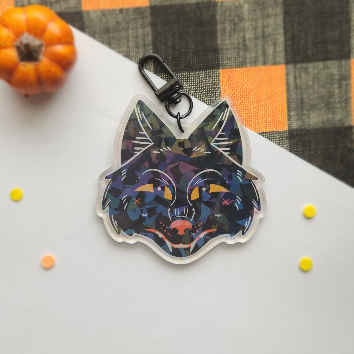 A die-cut acrylic keychain featuring a stylized black wolf face with a colorful geometric mosaic pattern, complete with a black metal clasp, displayed on a white surface with a small orange pumpkin and a plaid fabric background.