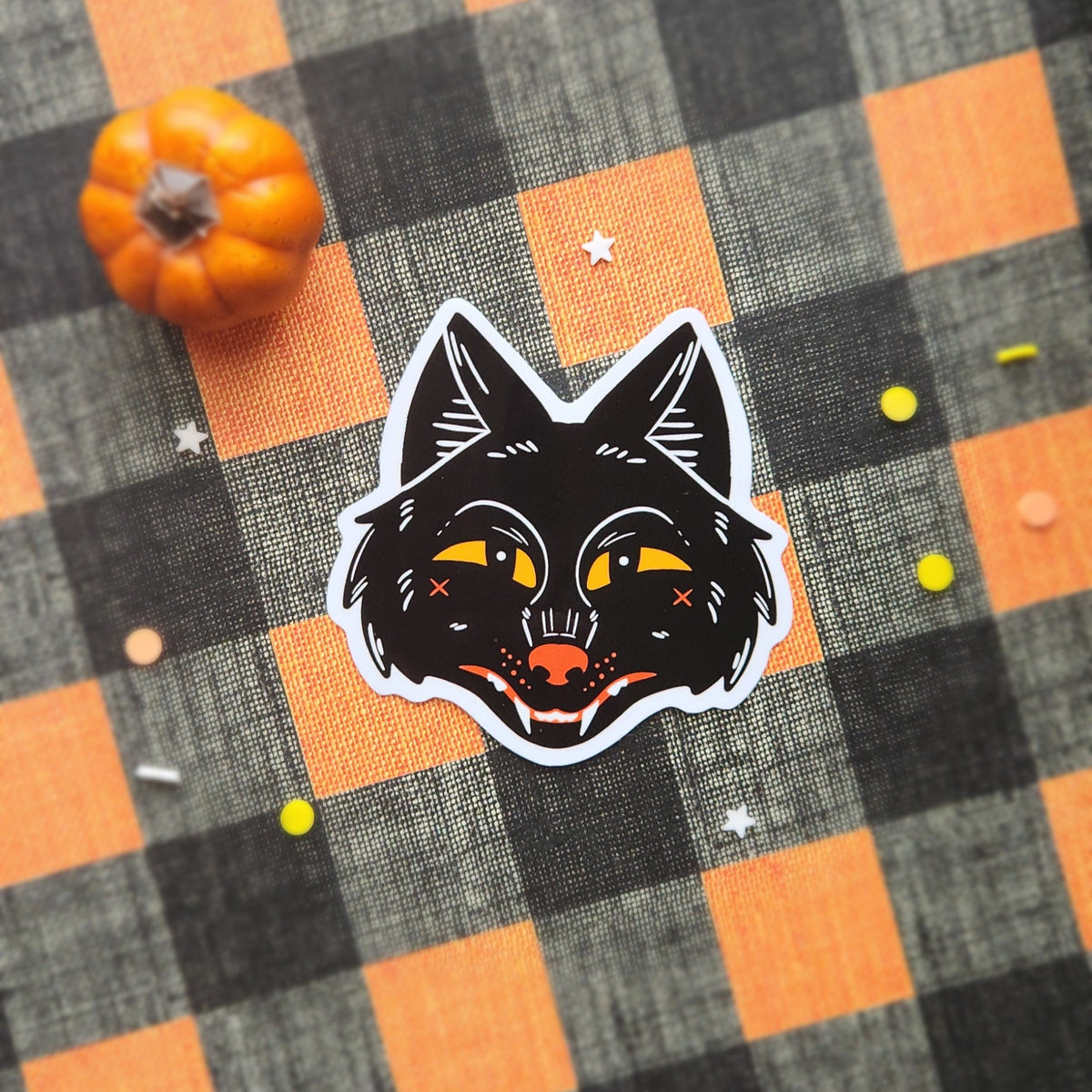 A die-cut sticker featuring a stylized black wolf face with yellow eyes and an orange nose, placed on an orange and black buffalo plaid fabric background with a small orange pumpkin prop.