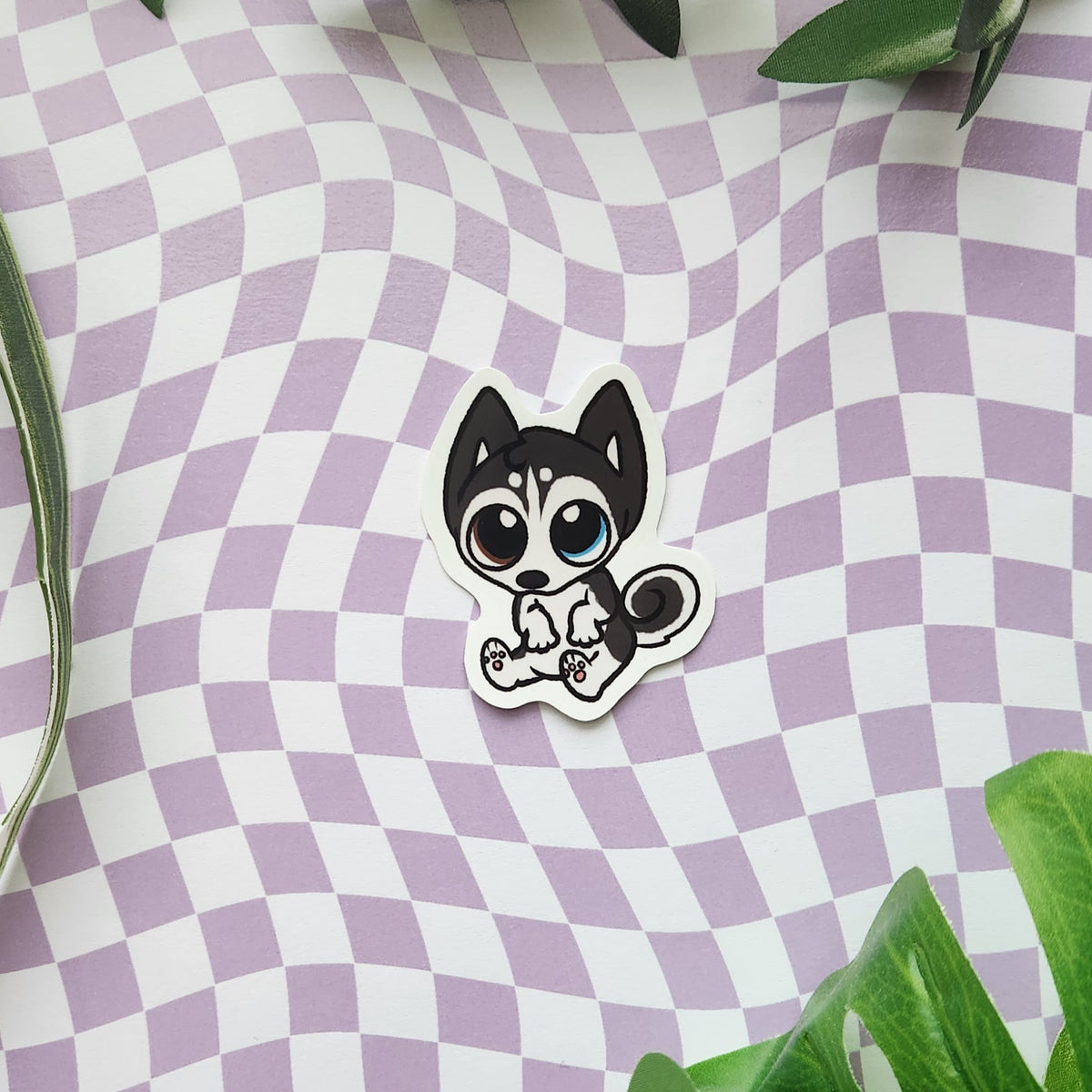 A die-cut sticker featuring a cute cartoon husky puppy, placed on a lavender and white wavy checkerboard patterned background with green leaves in the corners.