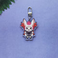 A chibi anime keychain featuring a white fox-like character with pink hair and orange-red wings, attached to a silver metal clasp, displayed on a textured lavender background with a green leaf in the corner.