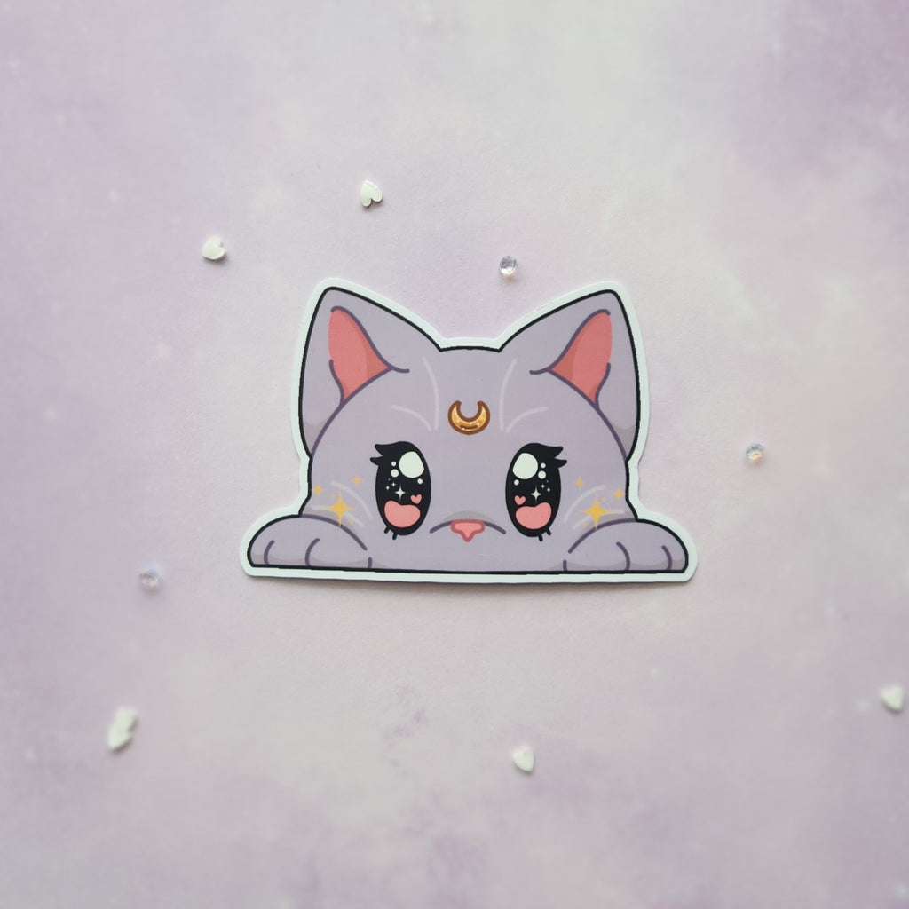 A die-cut sticker of a cute, lilac cat peeking over a white border, displayed on a light purple textured background with small scattered rhinestones.