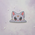 A die-cut sticker of a cute, lilac cat peeking over a white border, displayed on a light purple textured background with small scattered rhinestones.