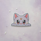 A die-cut sticker of a cute, lilac cat peeking over a white border, displayed on a light purple textured background with small scattered rhinestones.