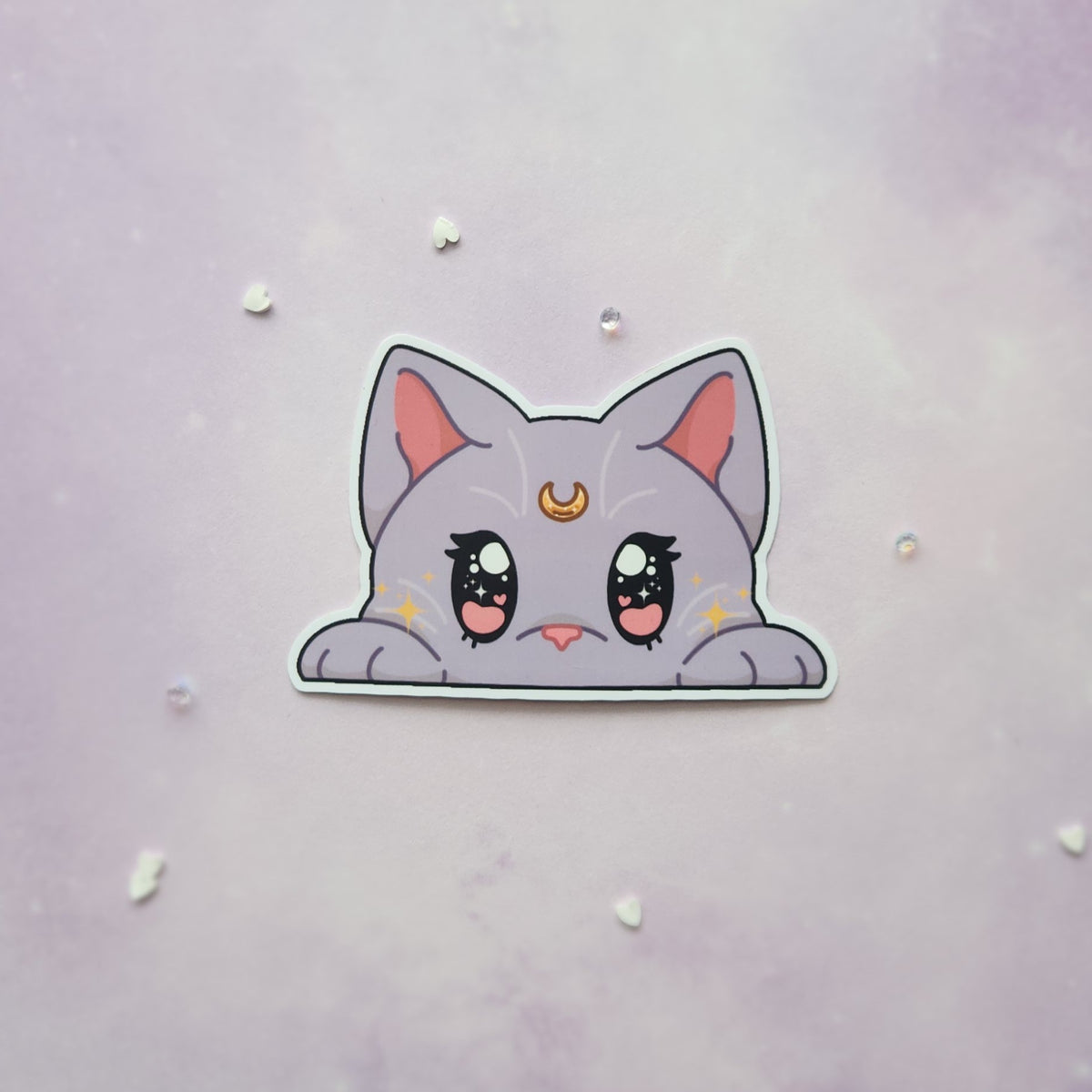 A die-cut sticker of a cute, lilac cat peeking over a white border, displayed on a light purple textured background with small scattered rhinestones.