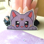A die-cut sticker featuring a cute purple cartoon cat face with pink inner ears, large expressive eyes, and a small gold crescent moon on its forehead, displayed in front of pink crystal points.