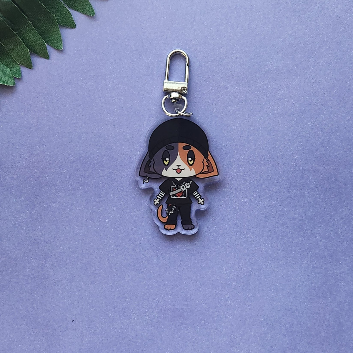 A small acrylic keychain charm featuring a stylized cartoon cat character in black clothing, attached to a silver lobster clasp, photographed on a soft lavender background with a small green leaf at the top left.