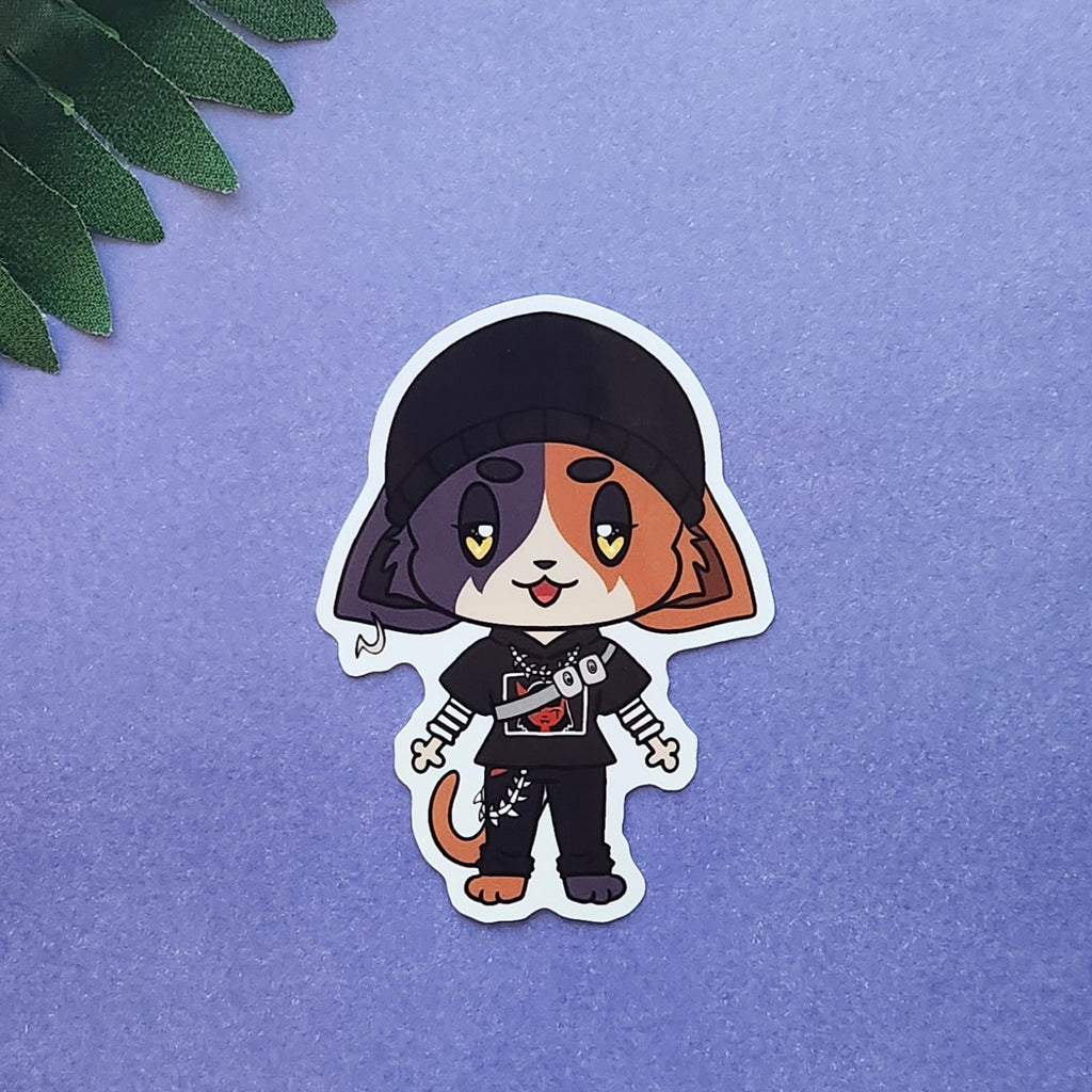 A die-cut sticker featuring a chibi-style cartoon cat character wearing a black cap, black outfit, and holding a small knife, placed on a textured purple surface with green leaves in the top-left corner.