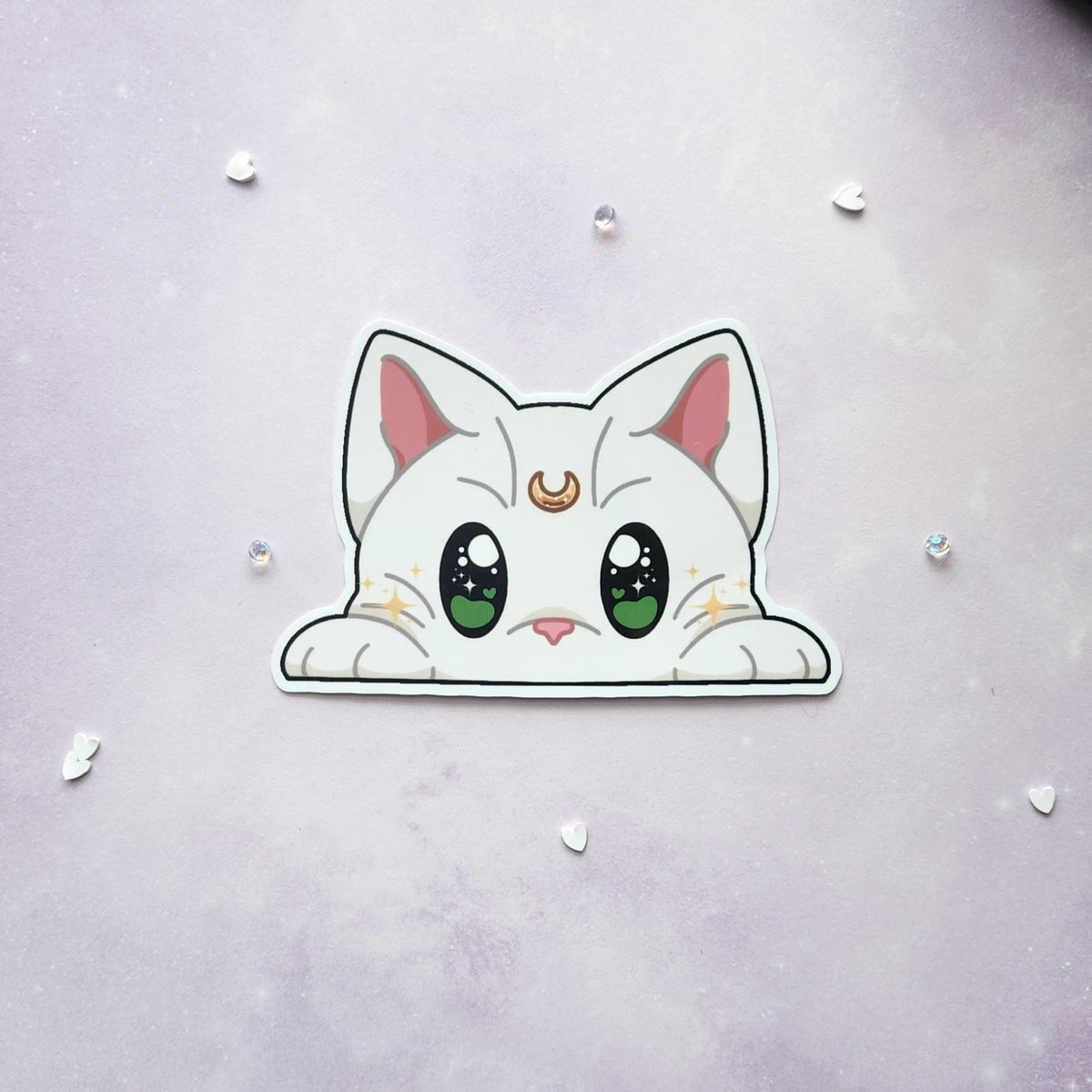 A cute cartoon cat sticker with large green eyes, pink inner ears, and a gold crescent moon on its forehead, shown on a soft lavender background with small scattered rhinestones.