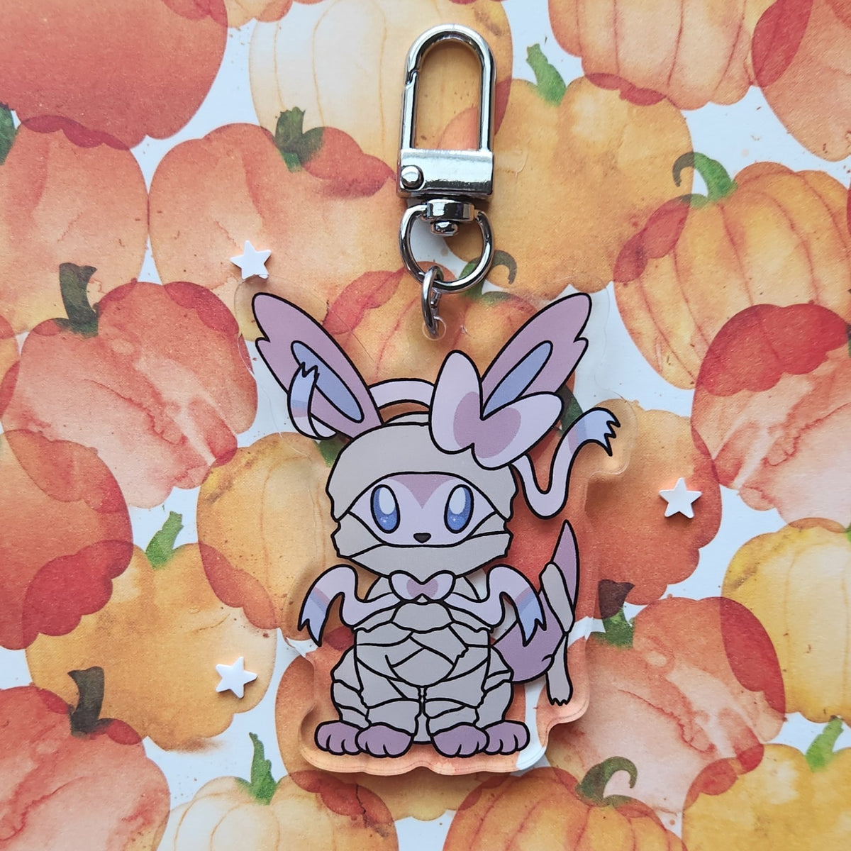 A cute fox keychain charm in a mummy costume, featuring a silver metal lobster clasp. The charm is made of layered acrylic with a light purple and gray mummy design, set against a watercolor pumpkin patterned background.