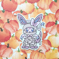 A die-cut vinyl sticker of a cute, mummy-wrapped fox with large ears and blue eyes, displayed on a watercolor pumpkin patterned background.