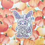 A die-cut vinyl sticker of a cute, mummy-wrapped fox with large ears and blue eyes, displayed on a watercolor pumpkin patterned background.