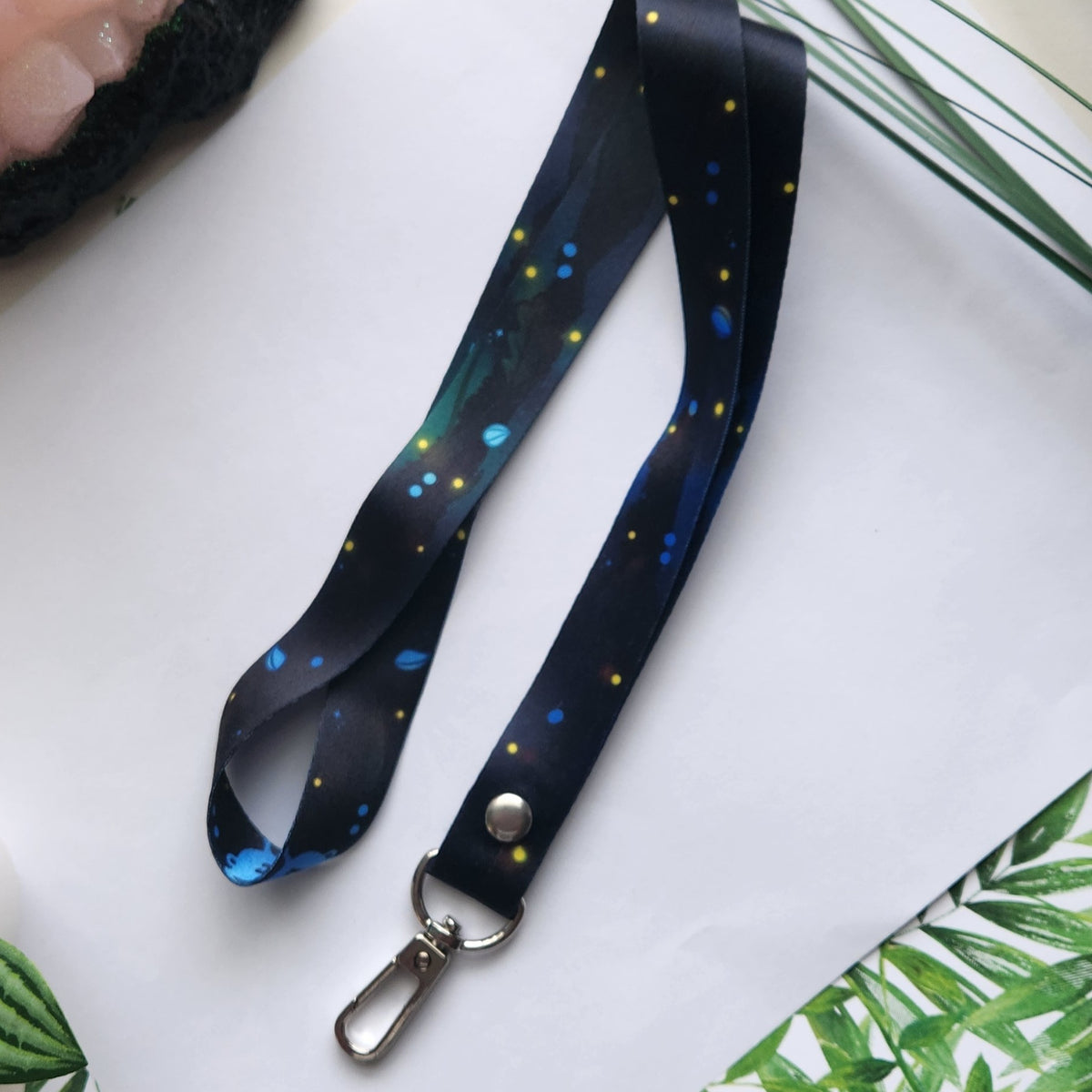 A black lanyard strap with a floral print of blue, green, and yellow flowers, featuring a silver metal snap button and lobster clasp, laid out on a white surface with decorative green leaves.