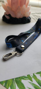 A black fabric lanyard with a blue, yellow, and white floral pattern, featuring a silver metal swivel clasp and a silver rivet, displayed on a light gray surface.