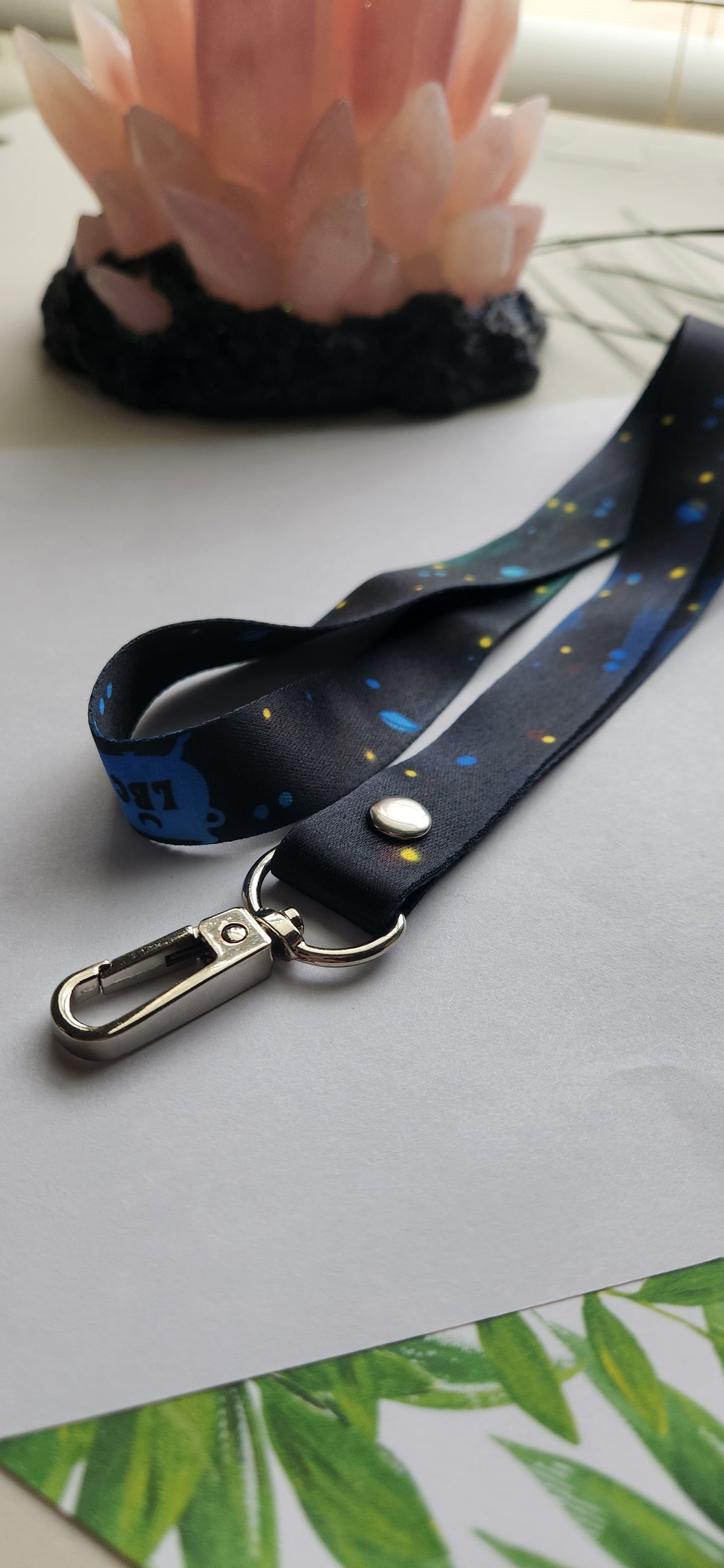 A black fabric lanyard with a blue, yellow, and white floral pattern, featuring a silver metal swivel clasp and a silver rivet, displayed on a light gray surface.
