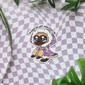 A die-cut sticker featuring a crying cartoon cat in a purple floral cloak with the text 'PLEASE DO NOT PERCEIVE ME' arched above its head, displayed on a lavender and white checkered surface with green leaves.