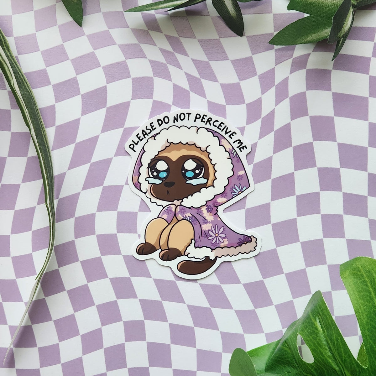 A die-cut sticker featuring a crying cartoon cat in a purple floral cloak with the text 'PLEASE DO NOT PERCEIVE ME' arched above its head, displayed on a lavender and white checkered surface with green leaves.