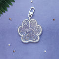 A paw-shaped acrylic keychain charm with a silver clasp, decorated with white crescent moons, stars, and small hearts filled with glitter, set against a textured purple background with scattered star confetti.
