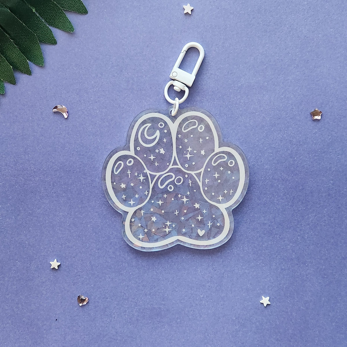 A paw-shaped acrylic keychain charm with a silver clasp, decorated with white crescent moons, stars, and small hearts filled with glitter, set against a textured purple background with scattered star confetti.