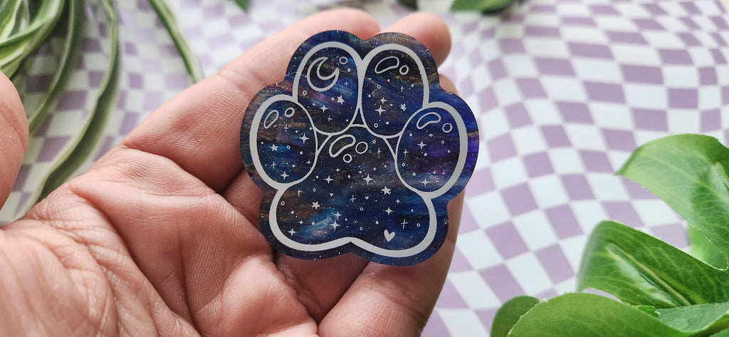 A hand holding a paw-shaped resin charm with a blue and purple galaxy pattern, set against a purple and white checkered background with green leaves.