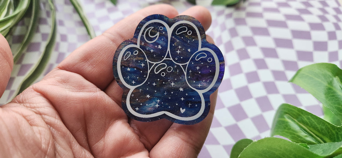 A hand holding a paw-shaped resin charm with a blue and purple galaxy pattern, set against a purple and white checkered background with green leaves.