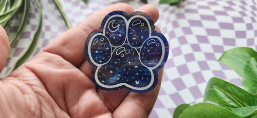 A hand holding a paw-shaped resin charm with a blue and purple galaxy pattern, set against a purple and white checkered background with green leaves.
