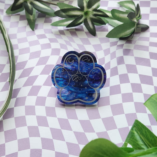 A paw-shaped resin pin with a blue galaxy pattern and white swirl details, displayed on a lavender and white checkered surface with green leaves.