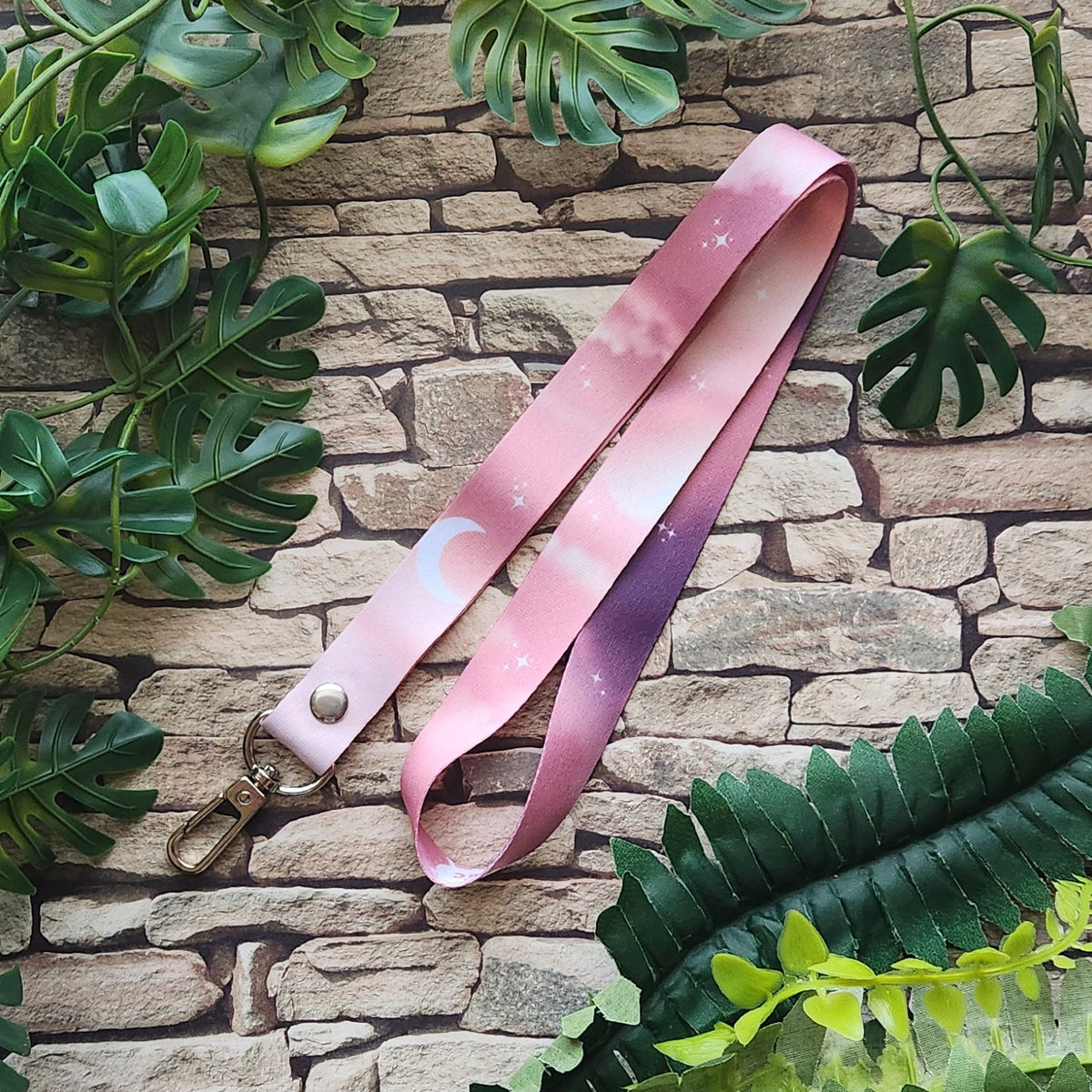 A pink gradient lanyard with a celestial print of stars and a crescent moon, featuring a gold-tone metal snap and clasp, laid out on a rustic stone wall with green foliage.