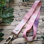 A pink gradient lanyard strap with a crescent moon and star pattern, attached to a silver metal lobster clasp, displayed on a stone background with green leaves.