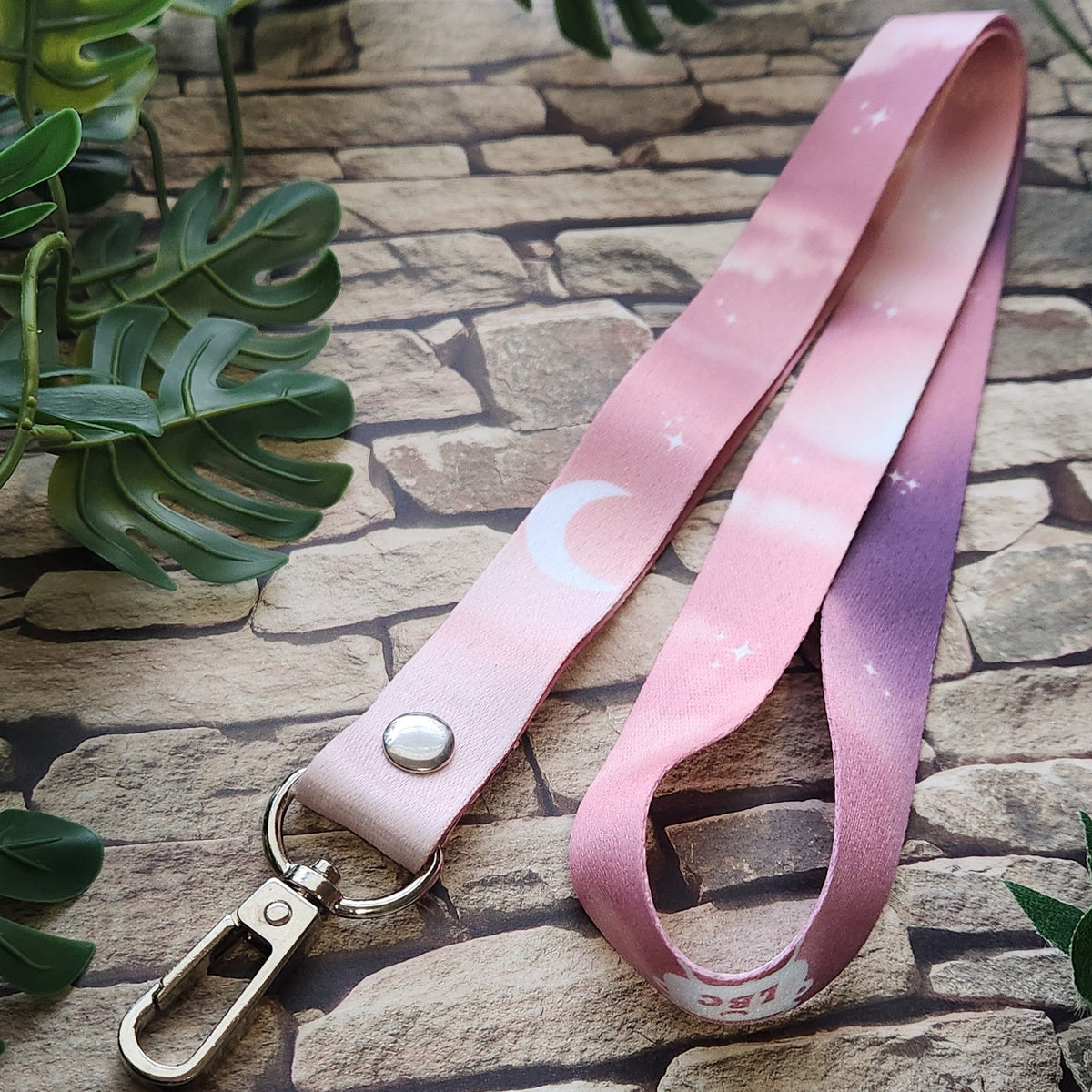 A pink gradient lanyard strap with a crescent moon and star pattern, attached to a silver metal lobster clasp, displayed on a stone background with green leaves.