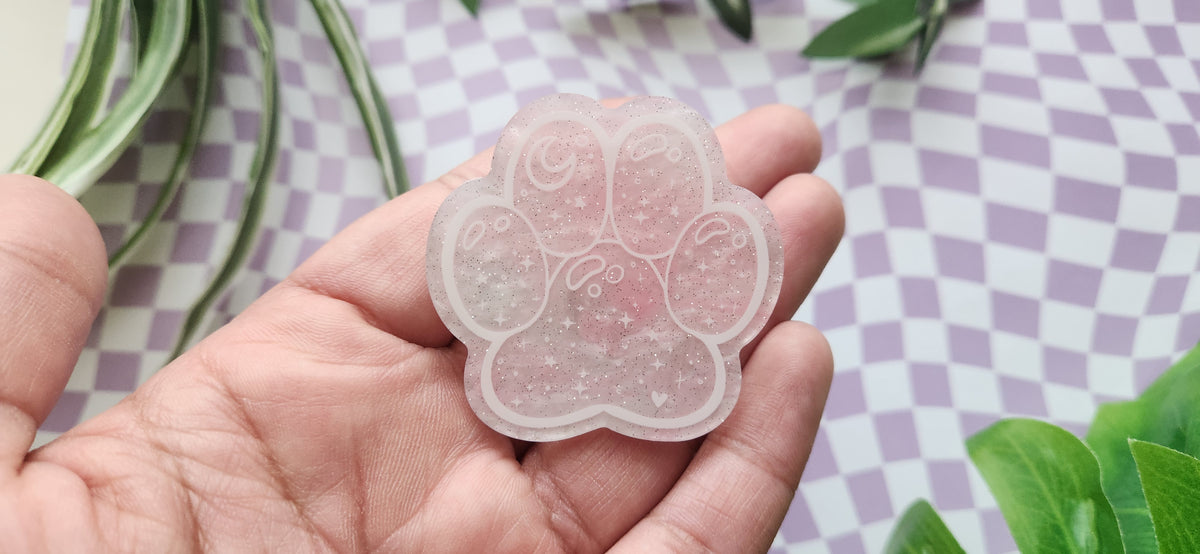 A translucent pink paw-shaped resin charm or pendant held in a person's hand, featuring a glittery texture and a small raised paw motif in the center, set against a purple and white checkered background with green leaves.