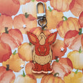 A cute acrylic keychain charm shaped like a pumpkin fox with large ears, attached to a silver metal clasp, set against a watercolor pumpkin patterned background.