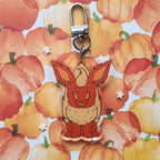 A cute acrylic keychain charm shaped like a pumpkin fox with large ears, attached to a silver metal clasp, set against a watercolor pumpkin patterned background.