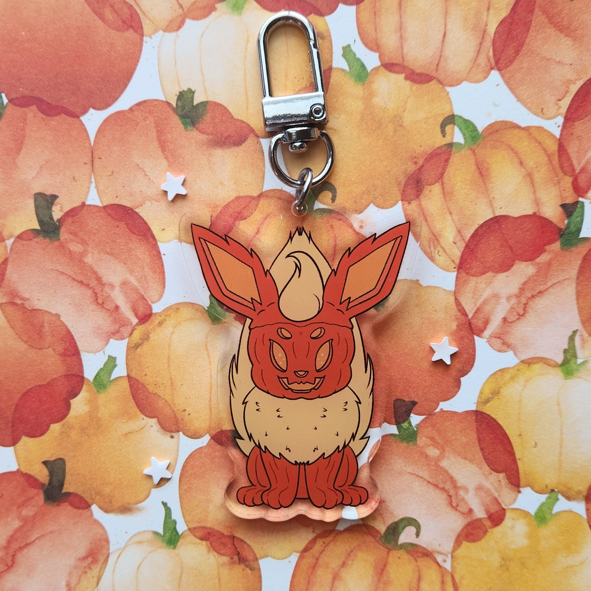 A cute acrylic keychain charm shaped like a pumpkin fox with large ears, attached to a silver metal clasp, set against a watercolor pumpkin patterned background.