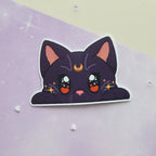 A die-cut sticker featuring a cute, kawaii-style black cat peeking up with large expressive eyes, orange cheeks, a gold crescent moon on its forehead, and sparkles, shown on a pastel purple background with small scattered star and heart embellishments.