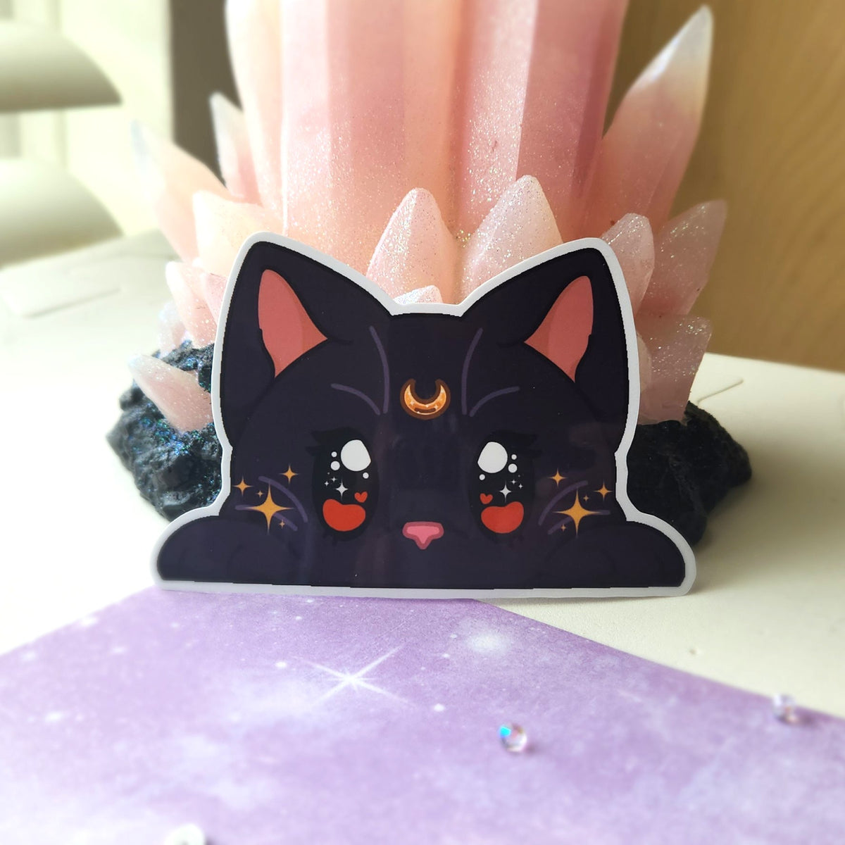 A die-cut sticker of a cute black cat with pink inner ears, white eyes, and a gold crescent moon on its forehead, displayed in front of a pink crystal cluster.