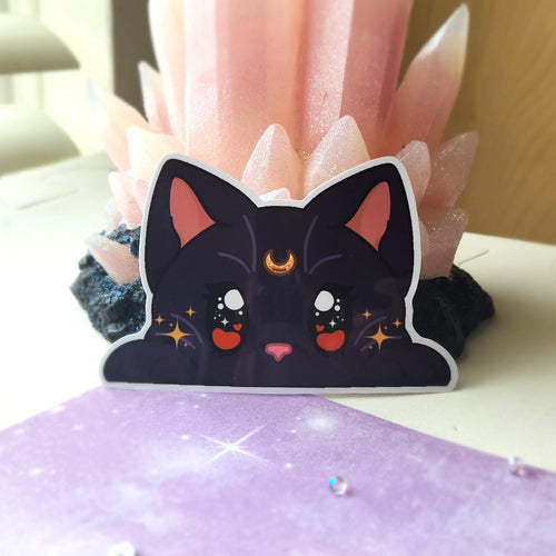 A die-cut sticker of a cute black cat with pink inner ears, white eyes, and a gold crescent moon on its forehead, displayed in front of a pink crystal cluster.