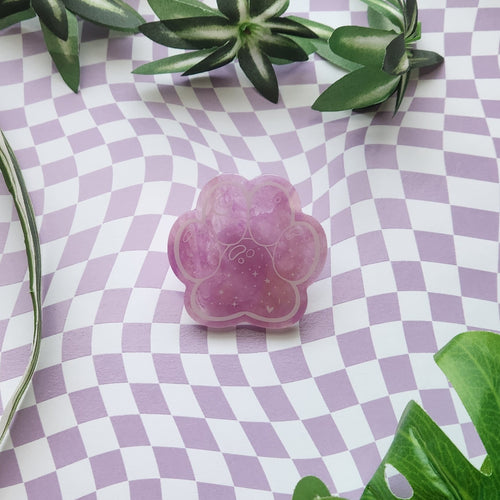 A pink paw-shaped resin charm with glittery purple inclusions, displayed on a lavender and white checkered background with green leaves.