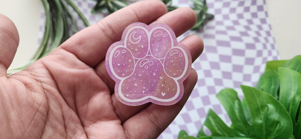 A hand holding a pink paw-shaped sticker with a glittery purple and gold design, set against a background of a purple-and-white checkered cloth and green leaves.
