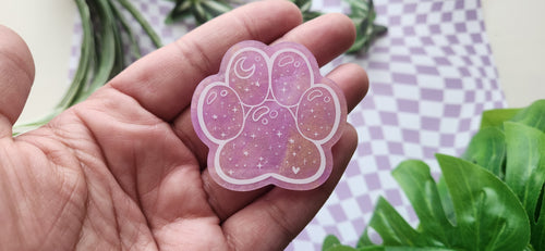 A hand holding a pink paw-shaped sticker with a glittery purple and gold design, set against a background of a purple-and-white checkered cloth and green leaves.