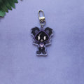 A small acrylic keychain charm featuring a stylized cartoon character in a dark purple and black outfit, attached to a silver metal clasp, photographed on a soft lavender background with a small green leaf visible in the top-left corner.