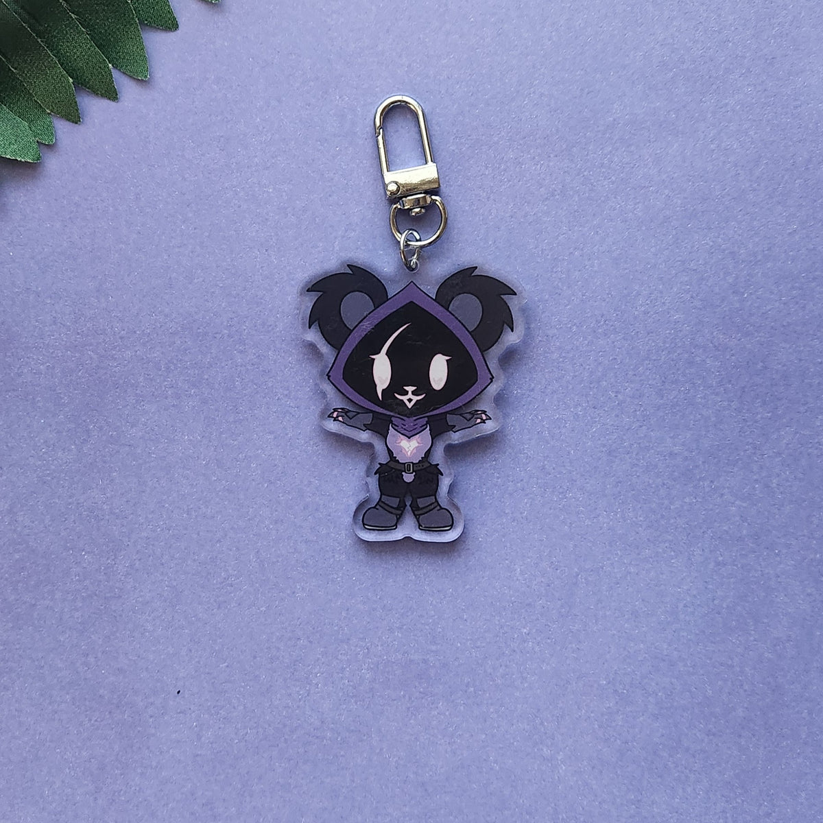 A small acrylic keychain charm featuring a stylized cartoon character in a dark purple and black outfit, attached to a silver metal clasp, photographed on a soft lavender background with a small green leaf visible in the top-left corner.