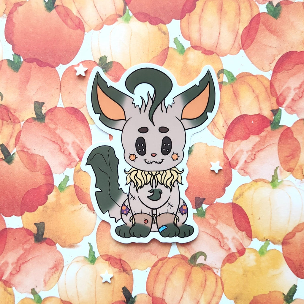 A die-cut sticker featuring a cute cartoon creature with large ears, a dark tail, and a ruffled blonde collar, placed on a background of watercolor pumpkins.