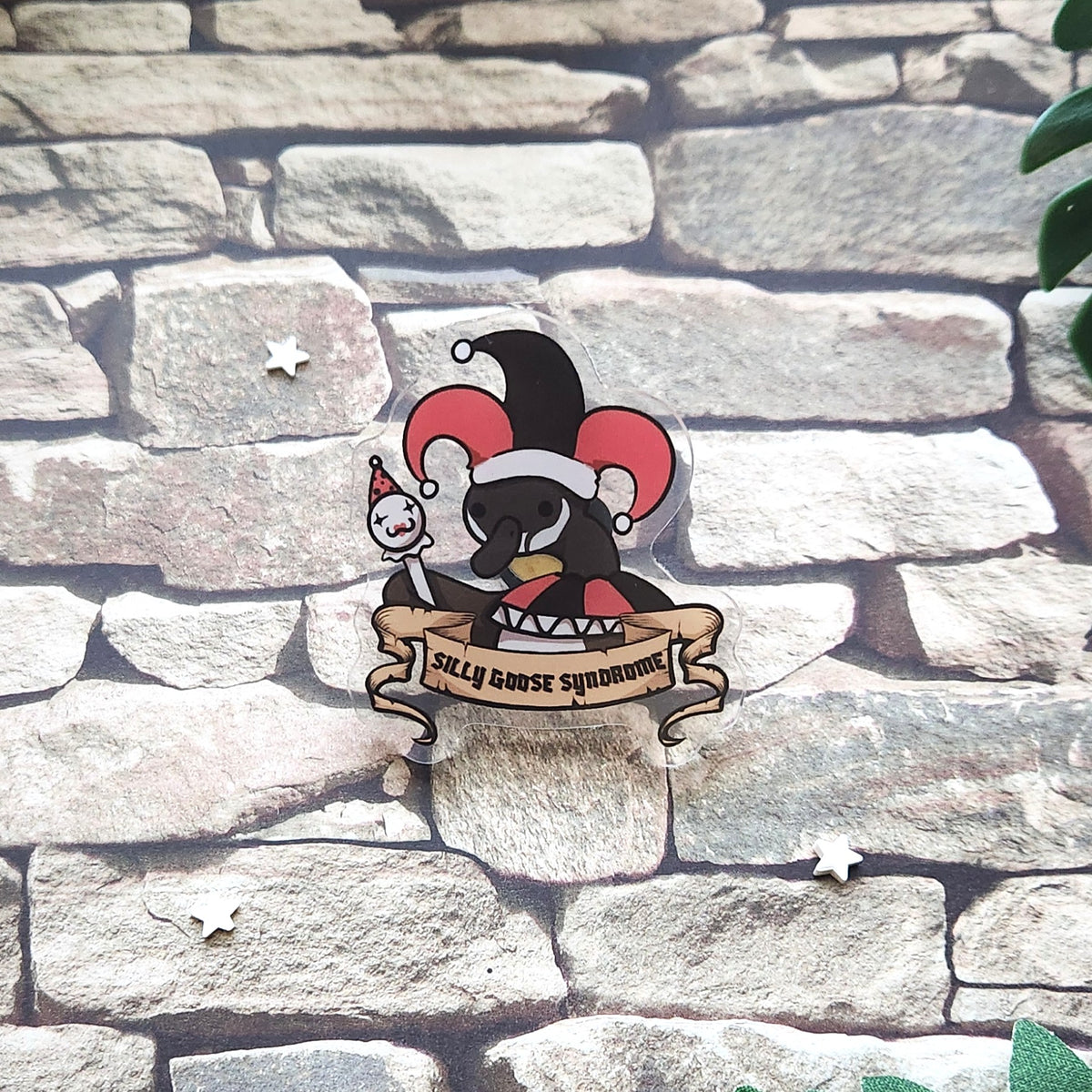 A pin featuring a cartoon goose in a jester hat with the text 'silly goose syndrome', laid on a stone backdrop.