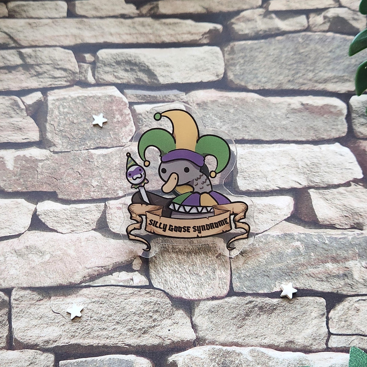 A pin featuring a cartoon goose in a jester hat with the text 'silly goose syndrome', laid on a stone backdrop.