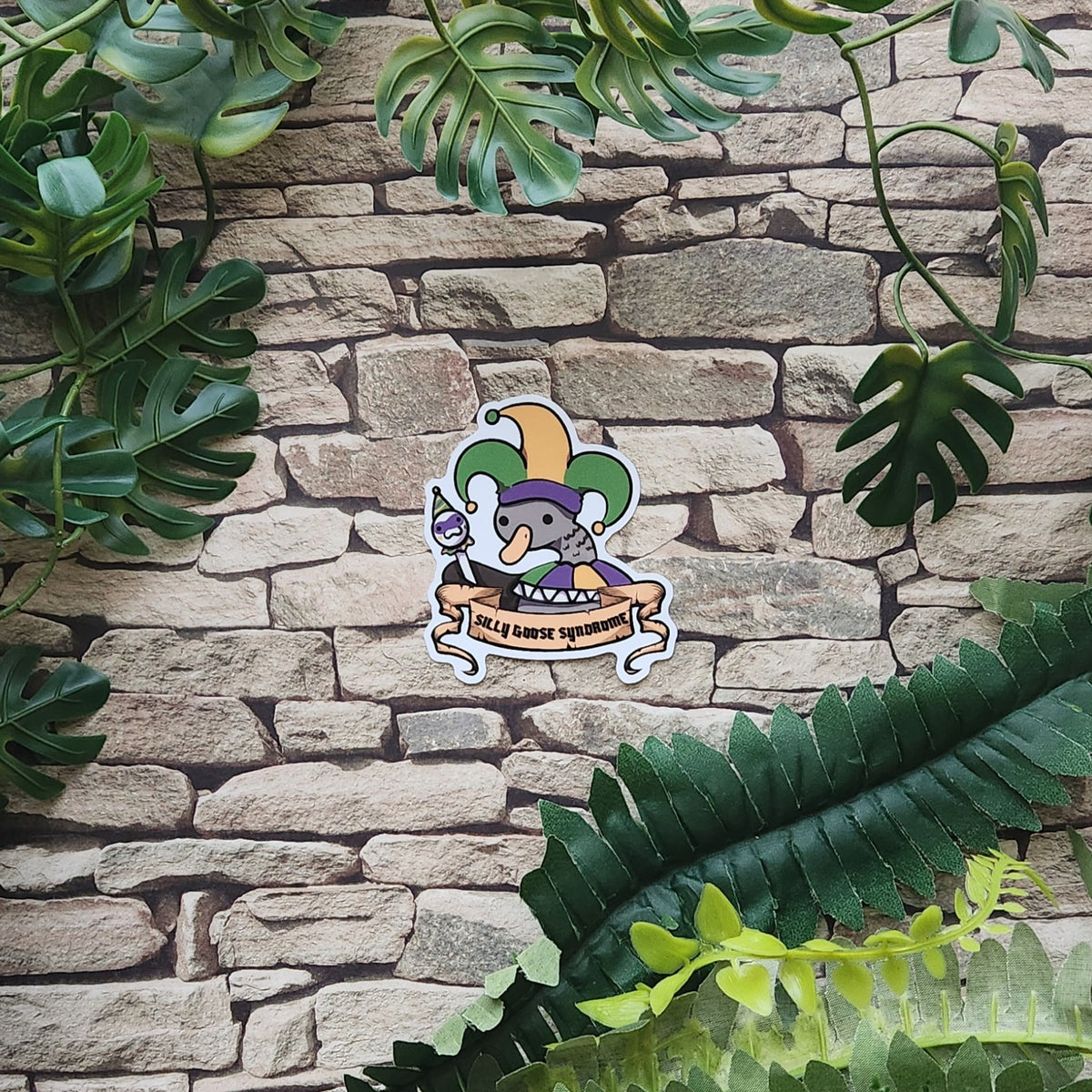 A die-cut sticker featuring a cartoon gray goose wearing a purple and green jester hat, with a banner reading 'Silly Goose Syndrome', displayed on a stone wall with green foliage.