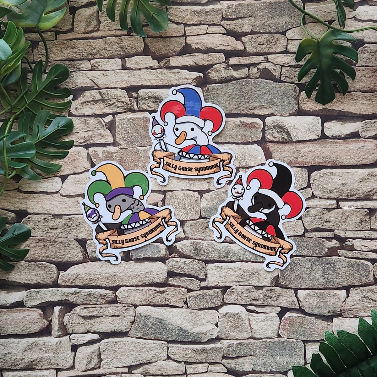 Three cartoon jester-themed stickers displayed on a stone wall background with green leaves framing the top and right sides. The stickers feature white jester characters with red, blue, green, and purple hats, holding banners that read 'Silly Goose Syndrome'.